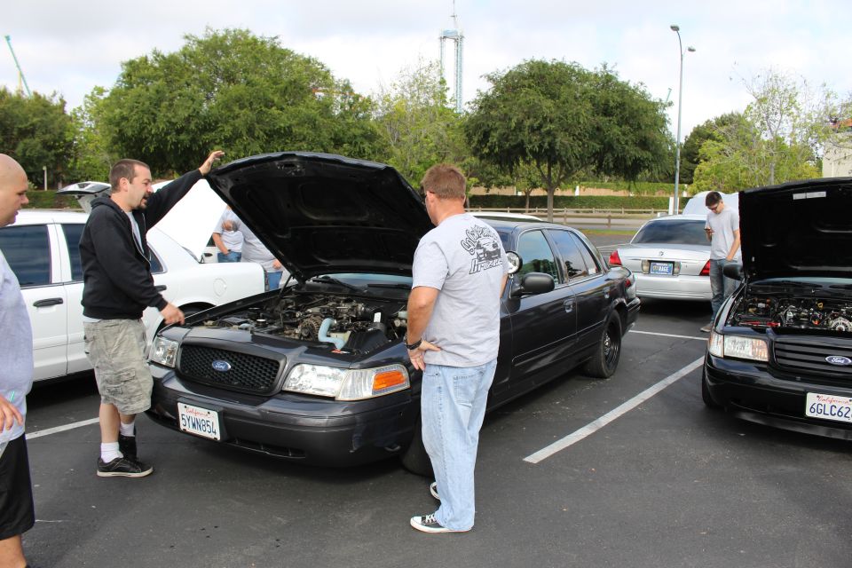 Men inspect a black Ford Crown Victoria with hood open at a car meet