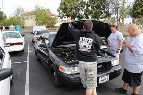 2003 Ford Crown Victoria - man in a graphic hoodie raises the hood of a black Ford Crown Victoria as others watch in a parking lot.