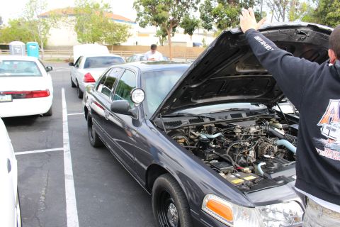 2003 Ford Crown Victoria - man lifts the hood of a dark gray Ford Crown Victoria with steel wheels at a parking-lot car meet.