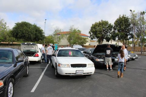 2003 Ford Crown Victoria - white Ford Crown Victoria with a show card on the dash anchors a busy parking-lot gathering of sedans.