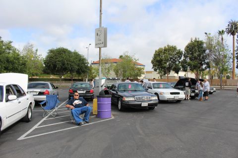 2003 Ford Crown Victoria - man sits in a chair beside black Panther sedans and a purple drum at an open parking-lot car meet.