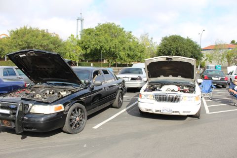 2003 Ford Crown Victoria - black Ford Crown Victoria with a brush guard sits beside a white sedan, both hoods raised, at a parking-lot meet.