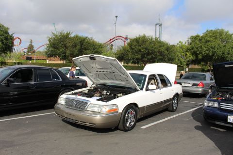 2003 Ford Crown Victoria - two-tone white and gold Mercury sedan sits with its hood up at a car show, roller coaster track in the background.