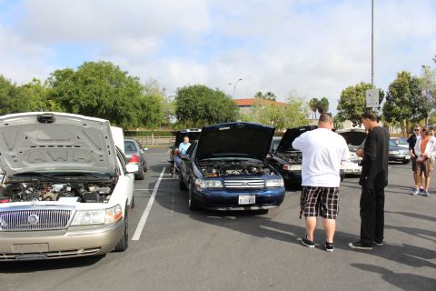 2003 Ford Crown Victoria - silver Mercury and a blue sedan show their engines while spectators in shorts stand alongside in a parking lot.