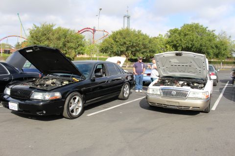 2003 Ford Crown Victoria - black Mercury sedan and a gold sedan sit with hoods raised at a car show, a roller coaster looming behind.