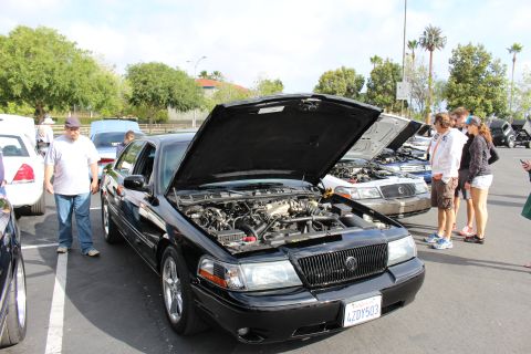 2003 Ford Crown Victoria - black Mercury sedan displays its V8 engine with hood up as spectators gather around in a sunny parking lot.