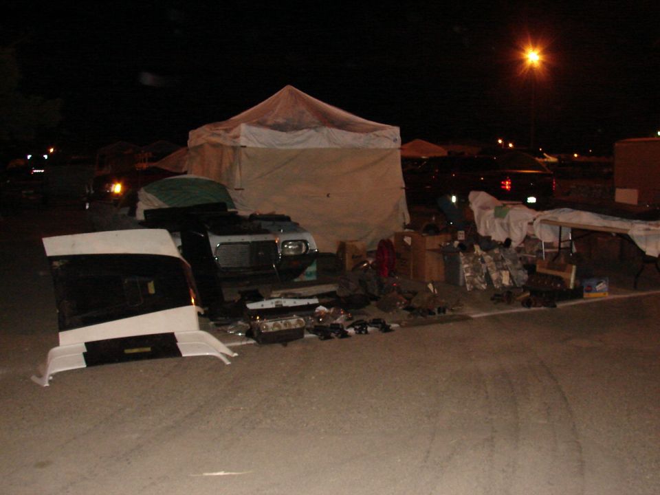 Car parts laid out on asphalt at night, with tents and a car in the background