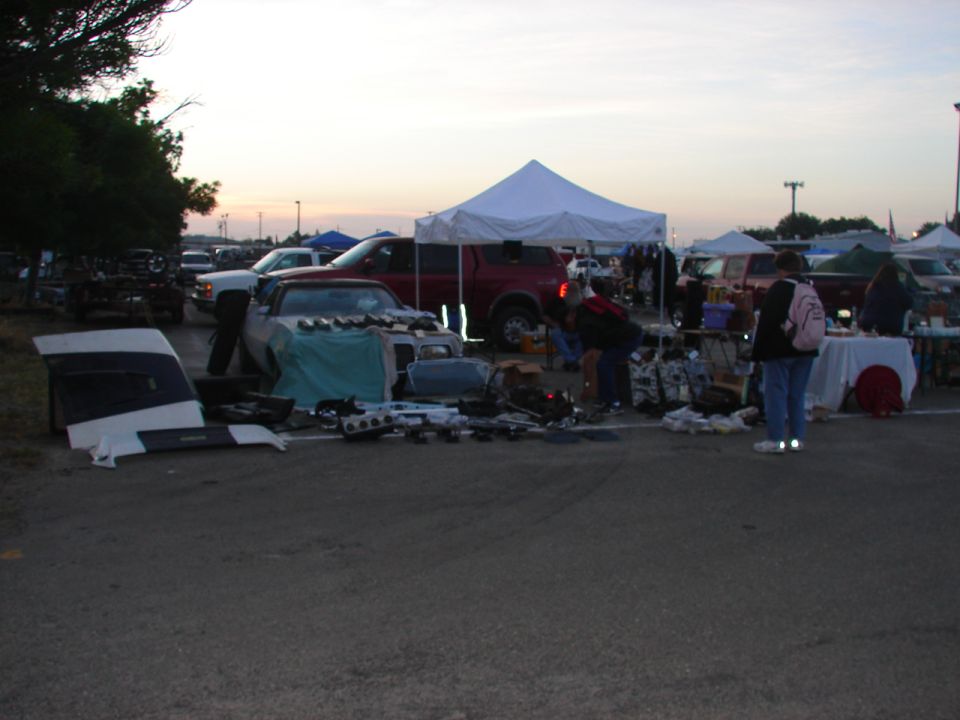 Car parts including a hood and engine components spread on asphalt at a swap meet