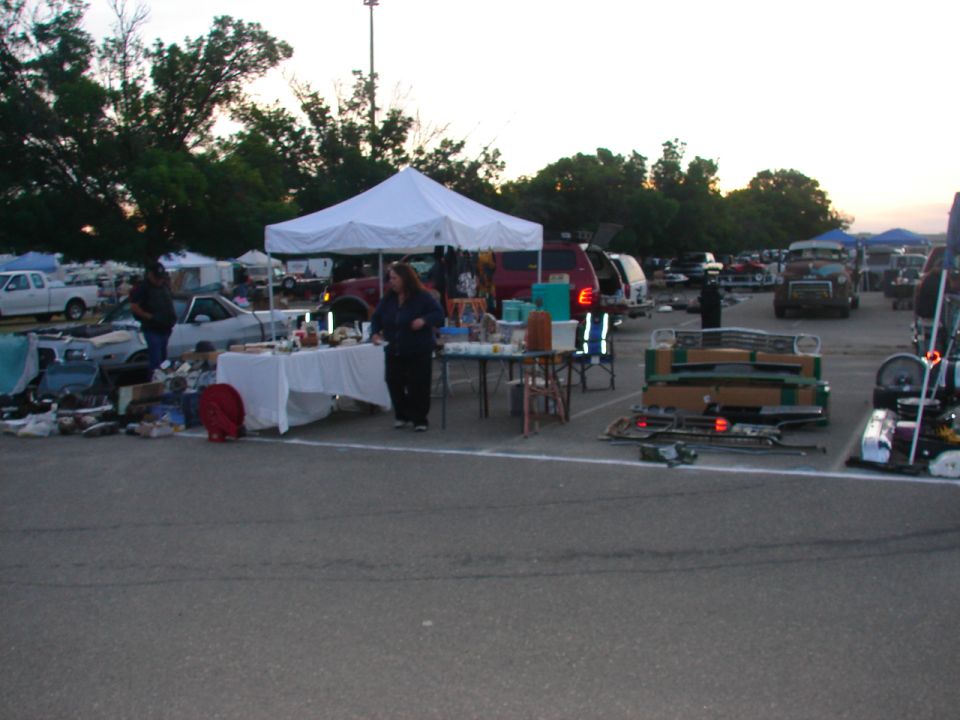Outdoor swap meet with tents, tables of goods, and vehicles parked in the background