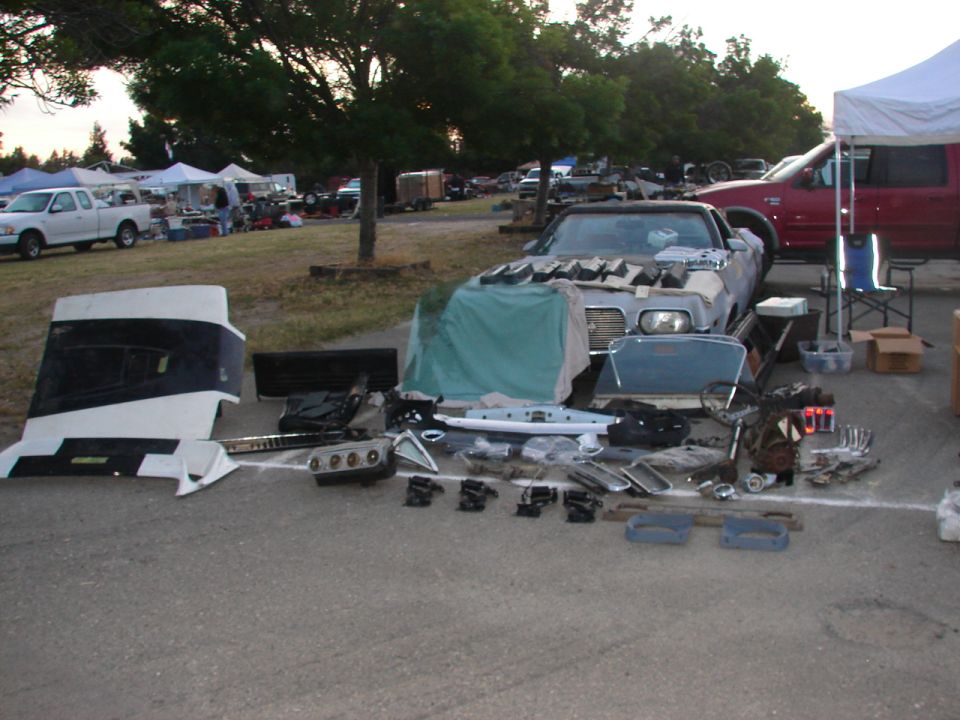 Car parts spread out on asphalt in front of a car covered with a tarp. Other vehicles and tents are in the background