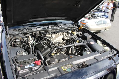 2003 Ford Crown Victoria - detailed V8 engine bay of a dark sedan sits exposed under a raised hood, a gold Mercury visible behind.