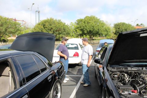 2003 Ford Crown Victoria - Two men in caps chat in a parking lot beside cars with open trunk and hood, a roller coaster behind them.