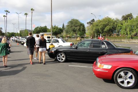 2003 Ford Crown Victoria - Spectators stroll past a black sedan with a flag and a red sedan in a parking-lot car show under palm trees.