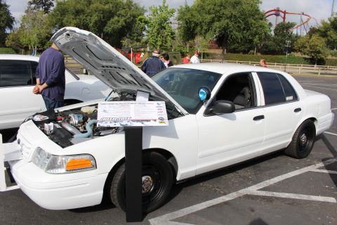 2003 Ford Crown Victoria - white Ford Crown Victoria displays its engine and a printed spec card on steel wheels at an outdoor car show.
