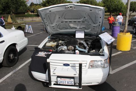 2003 Ford Crown Victoria - white Ford sedan with a push bar and 'GHST VIC' plate shows a supercharged engine bay at an outdoor car show.