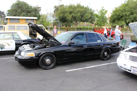 2003 Ford Crown Victoria - black Ford Crown Victoria sedan with steel wheels and open hood parked at an outdoor car show near a CHP-marked cruiser.