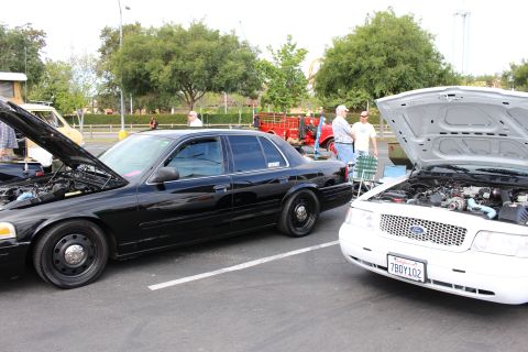 2003 Ford Crown Victoria - black Ford Crown Victoria sits beside a white Ford sedan with its hood up at an outdoor parking-lot car show.