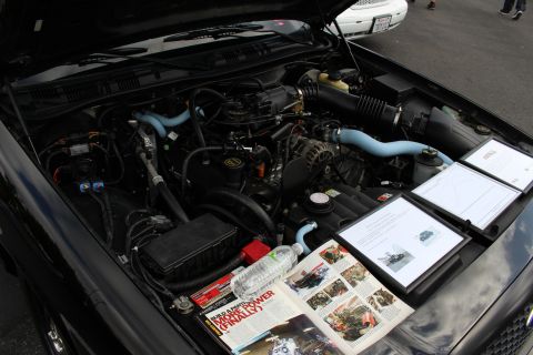2003 Ford Crown Victoria - Engine bay of a Ford sedan with blue silicone hoses, a clipboard display board, and an open car magazine resting on the fender.