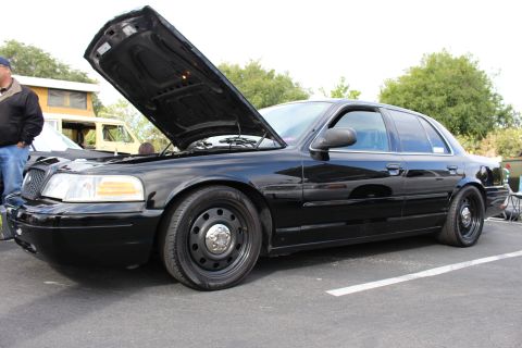 2003 Ford Crown Victoria - Side three-quarter view of a black Ford Crown Victoria with raised hood and black steel wheels at an outdoor car show.