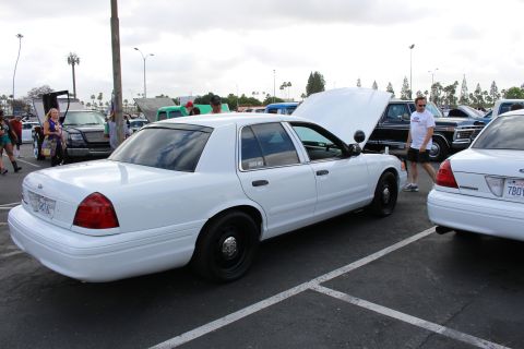 2003 Ford Crown Victoria - white Ford full-size sedan with steel wheels sits in a parking-lot car show, hood and trunk open under an overcast sky.
