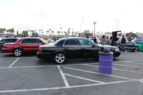 2003 Ford Crown Victoria - black full-size sedan on silver alloys sits in a striped parking lot next to a purple drum, with rows of show cars and palms behind.