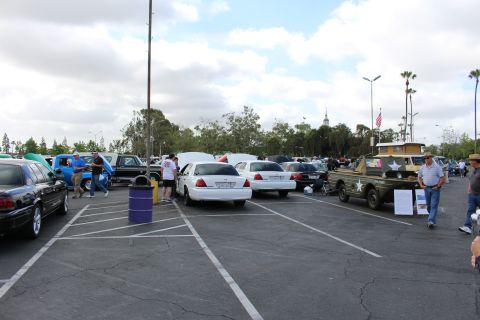 2003 Ford Crown Victoria - wide parking-lot car show scene with a white sedan center, a green military-style vehicle at right, attendees, and a flag in the distance.