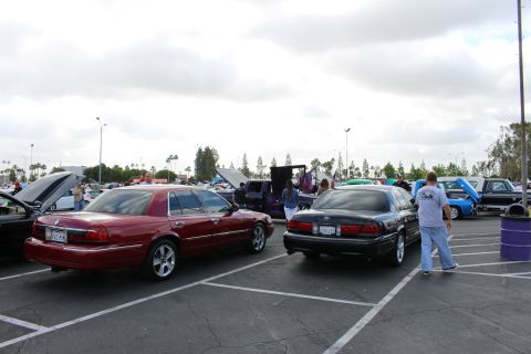 2003 Ford Crown Victoria - maroon sedan and a black sedan park back-to-back in a lot while a man in a gray shirt walks between rows of show cars.