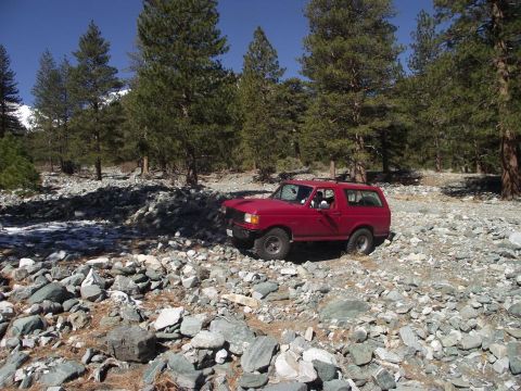 1988 Ford Bronco - A red SUV navigates a rocky, wooded area under a clear blue sky.