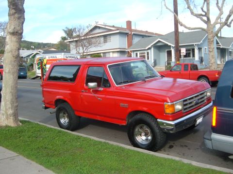 1988 Ford Bronco - A red Ford Bronco is parked curbside on a residential street with a U-Haul truck and another red pickup visible.