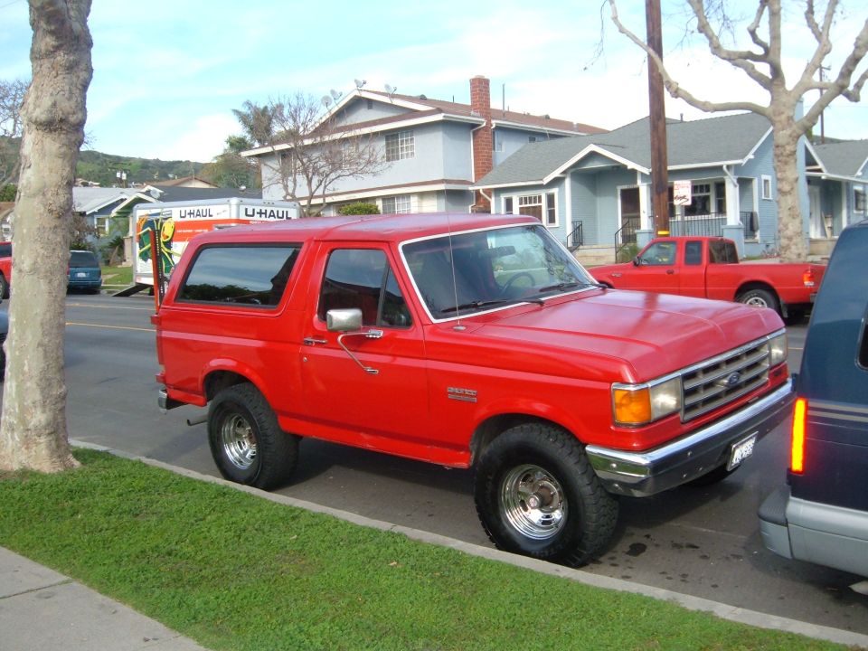 Red 1988 Ford Bronco parked on street, U-Haul truck and red pickup in background