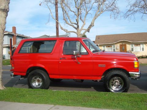 1988 Ford Bronco - A red Ford Bronco is parked on the side of the road with houses in the background.