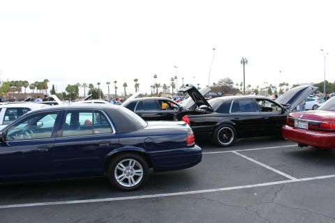 2003 Ford Crown Victoria - dark blue full-size sedan sits in the foreground of a packed parking-lot car show with several cars showing open hoods and trunks.