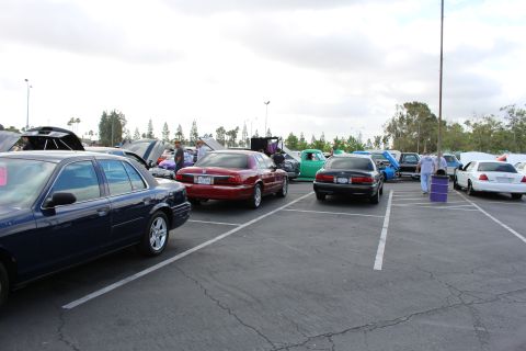 2003 Ford Crown Victoria - Navy, maroon and white full-size sedans park across several stalls of a parking-lot car show with people and a purple barrel behind.