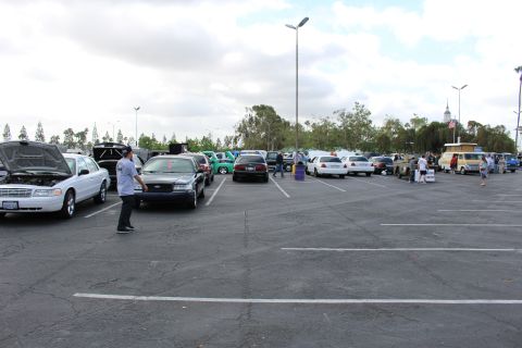 2003 Ford Crown Victoria - wide parking-lot car show view with a silver Ford sedan at left, scattered show cars and vendors, and a man walking across the asphalt.
