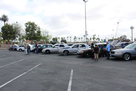 2003 Ford Crown Victoria - Silver and dark full-size sedans park at angles across a striped lot as small groups of attendees gather between the show cars.