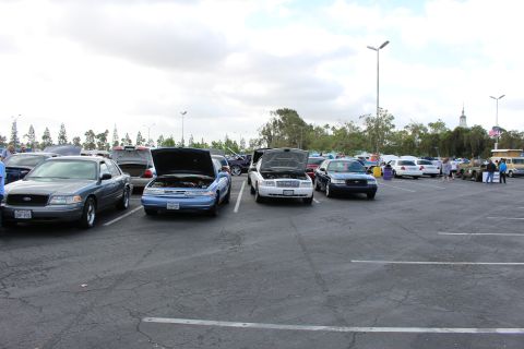 2003 Ford Crown Victoria - Gray, light-blue and white full-size sedans sit with hoods raised across a broad parking-lot car show with palms and a flag behind.