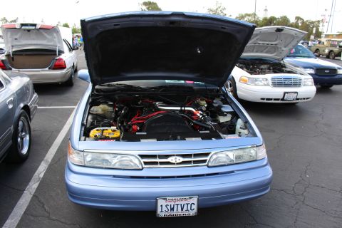 2003 Ford Crown Victoria - light-blue Ford full-size sedan shows its open hood and engine bay head-on, with a 1SWTVIC plate, amid other show cars.