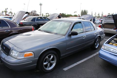 2003 Ford Crown Victoria - gray Ford full-size sedan on machined alloy wheels sits in front-three-quarter view among other cars with open hoods at a show.