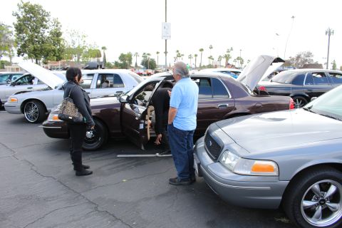 2003 Ford Crown Victoria - woman and a man in a blue shirt inspect a brown full-size sedan with its door open, flanked by silver Ford sedans at a car show.