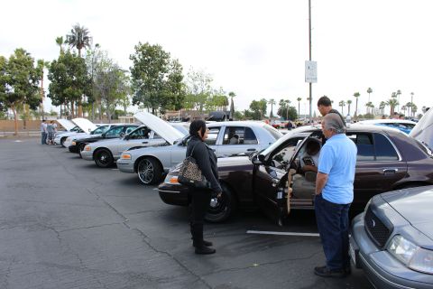 2003 Ford Crown Victoria - maroon Ford sedan sits with its driver door open as people inspect a row of cars at an outdoor parking-lot meet.