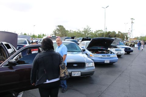 2003 Ford Crown Victoria - light-blue Ford sedan wearing a CHP PI1 plate is parked in a row of open-hood cars while a man in blue talks to a woman.