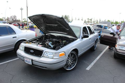 2003 Ford Crown Victoria - silver Ford sedan sits with its hood up showing the engine, riding on large multi-spoke chrome wheels at an outdoor meet.
