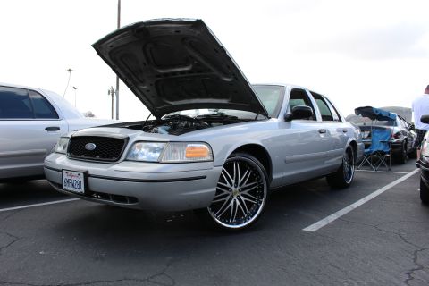 2003 Ford Crown Victoria - silver Ford sedan displays its open hood and large black-and-chrome multi-spoke wheels in a sunlit parking-lot car show.