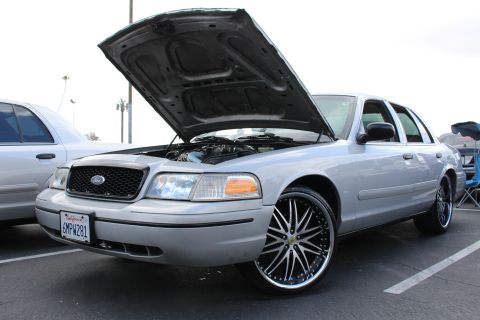 2003 Ford Crown Victoria - silver Ford sedan with raised hood and oversized chrome multi-spoke wheels parked in a marked lot under overcast sky.
