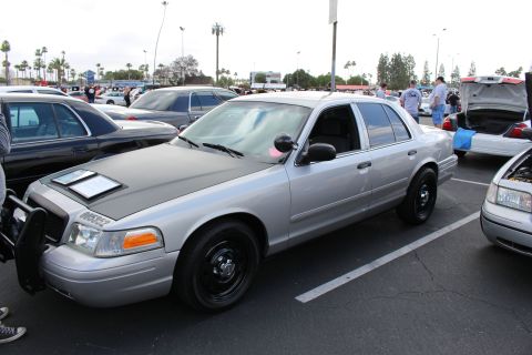 2003 Ford Crown Victoria - silver Ford sedan with black steel wheels and a matte-black hood section is parked among many cars at a packed outdoor meet.