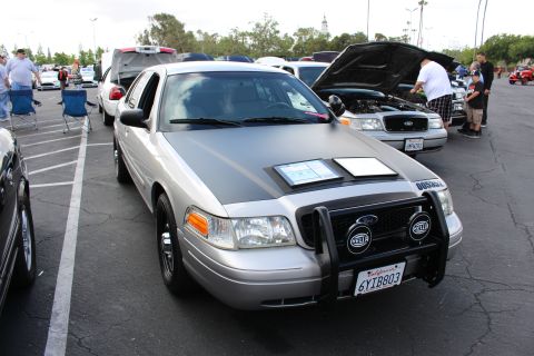 2003 Ford Crown Victoria - tan Ford sedan with a front push bar, Hella lights and a matte hood section is parked among show cars with spectators nearby.
