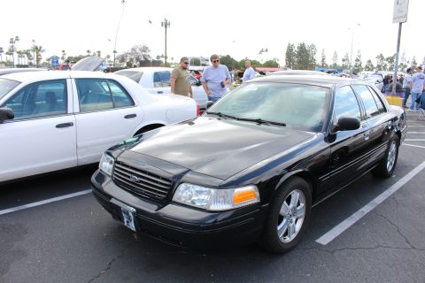 2003 Ford Crown Victoria - glossy black Ford sedan is parked next to a white sedan as two men stand behind it at a crowded outdoor car meet.