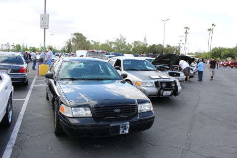 2003 Ford Crown Victoria - black Ford sedan heads a row of parked show cars, several with raised hoods, as people walk the lot under an overcast sky.