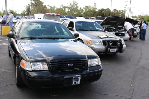 2003 Ford Crown Victoria - black Ford sedan sits ahead of a silver push-bar-equipped sedan in a long row of show cars as spectators browse the lot.