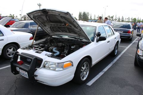 2003 Ford Crown Victoria - white Ford sedan with a front push bar and raised hood shows its engine while parked among many cars at a packed lot.